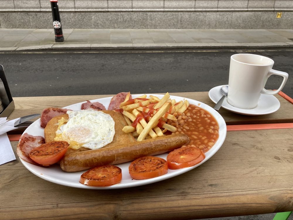 A breakfast plate of sausage, bacon, fried egg, grilled tomatoes, beans and chips with a cup of tea on an outdoor wooden table beside the pavement at JD Cafe